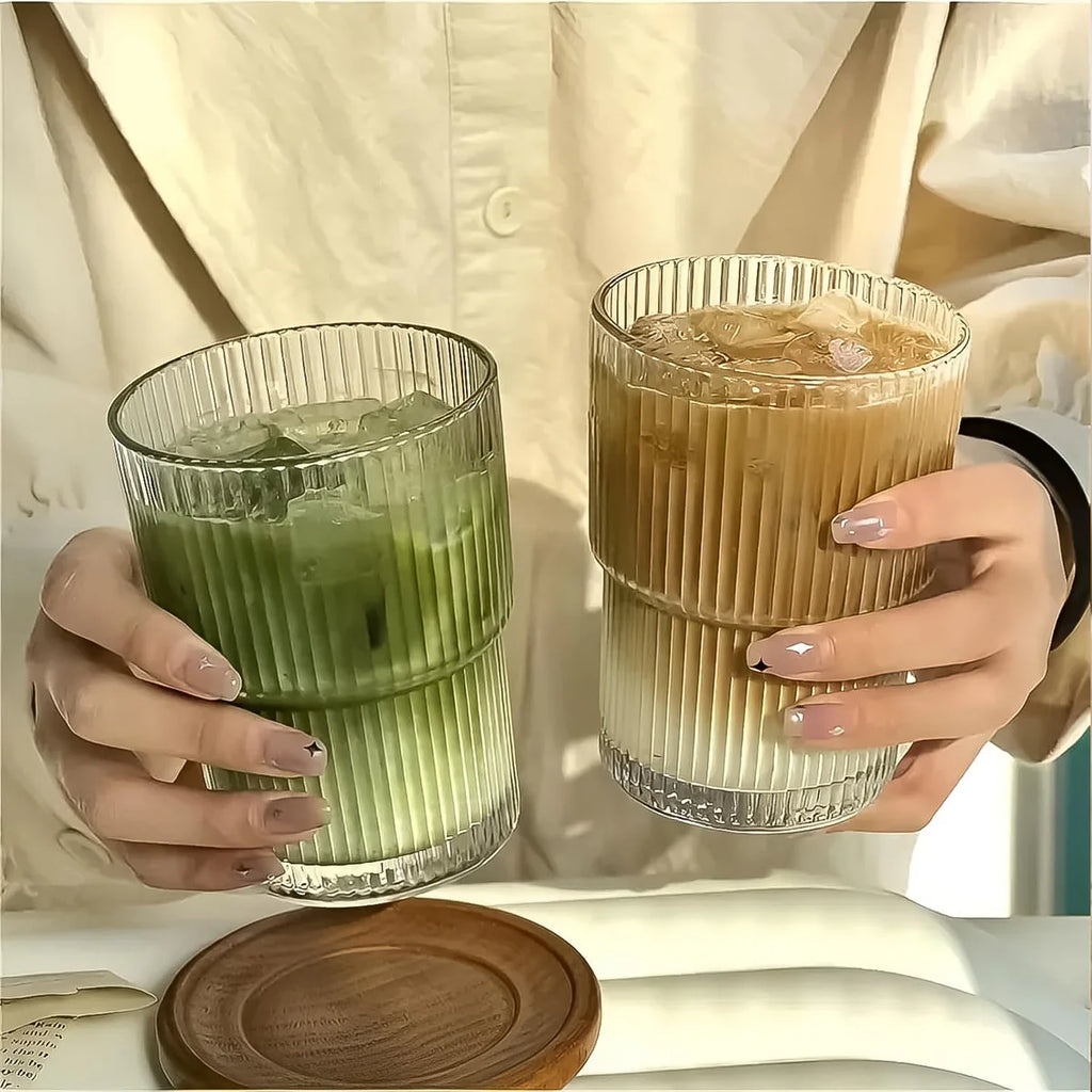 Two hands holding glasses of iced green and brown beverages on a light background